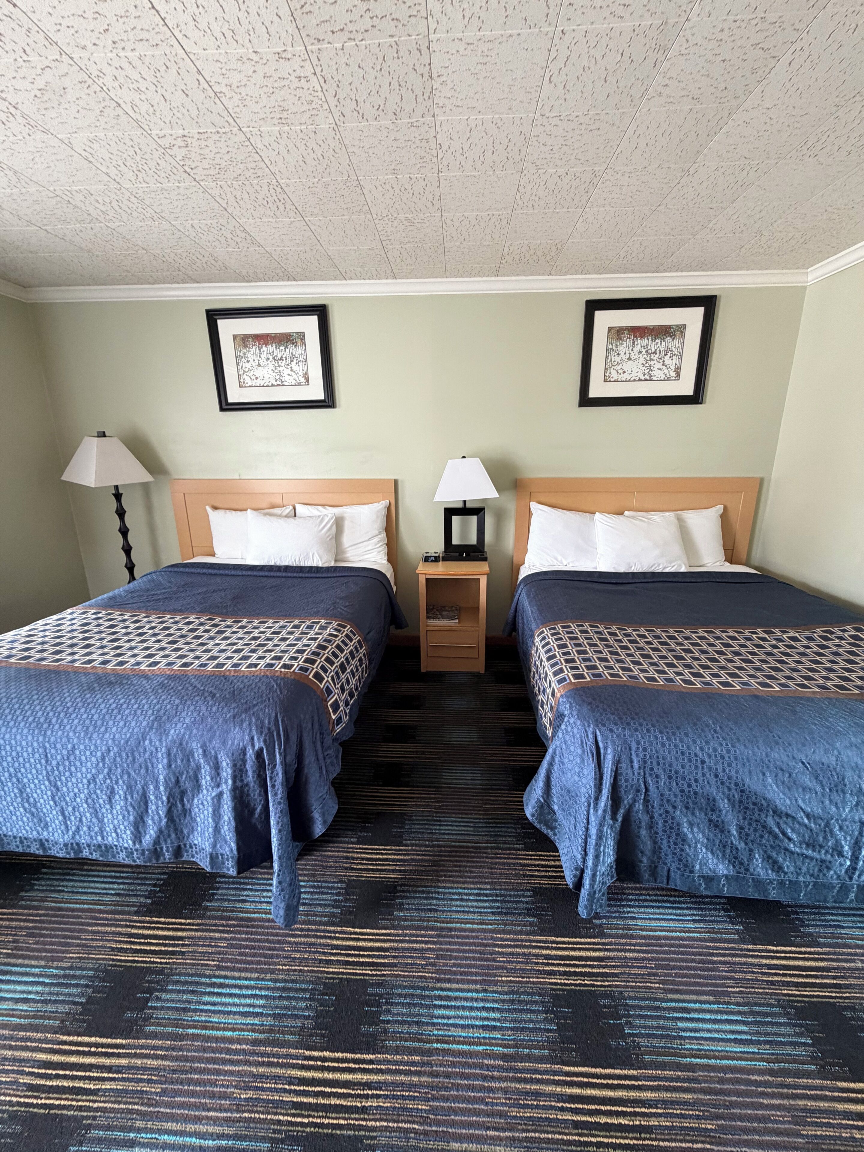 A hotel room with two beds, a nightstand, a lamp, and framed artwork on the wall.