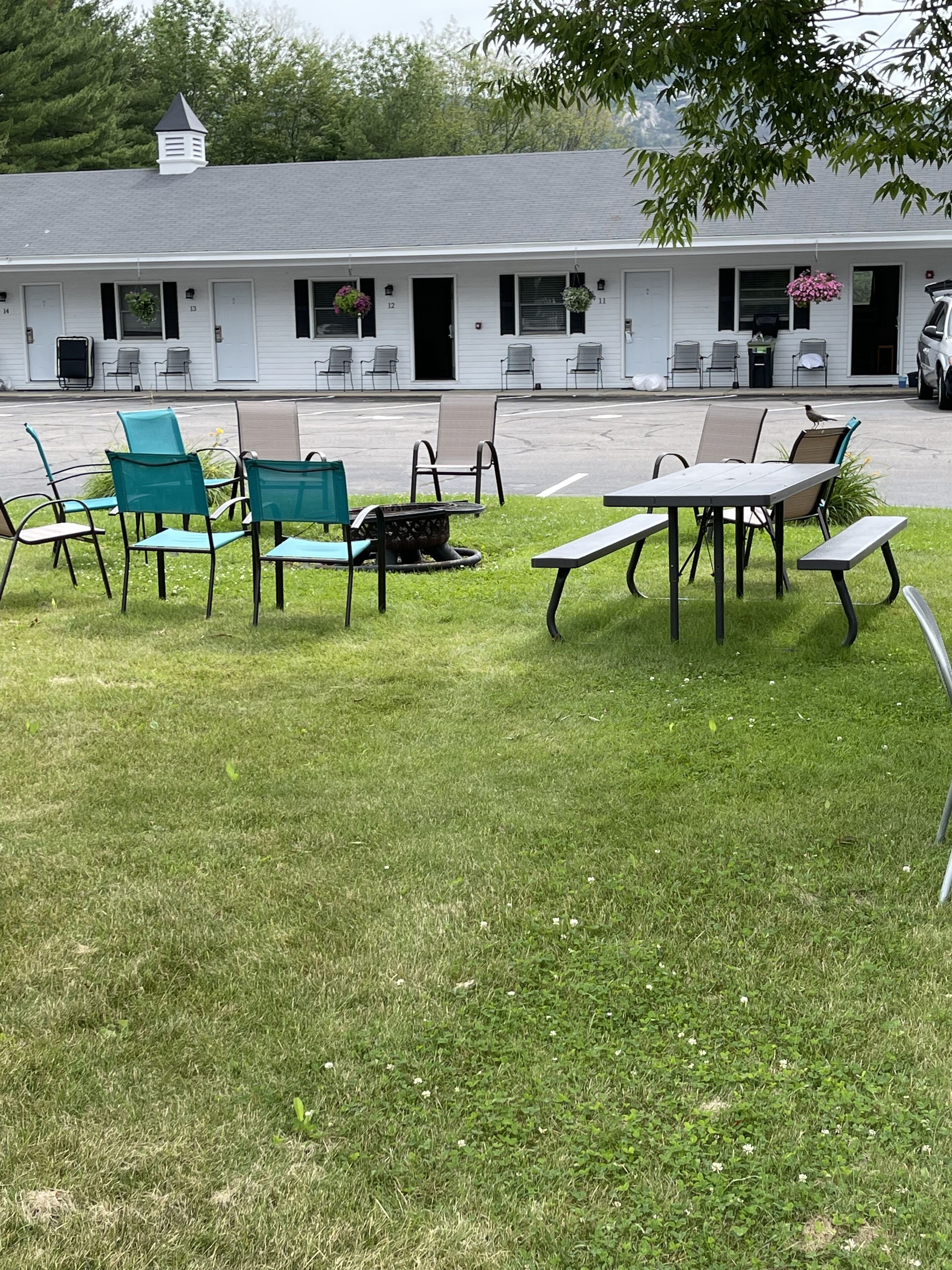 Un espace extérieur aménagé avec des chaises et des tables, un bâtiment dont les portes sont numérotées, et un parking en arrière-plan.