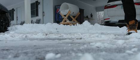 Una strada di accesso coperta di neve con un oggetto bianco di grandi dimensioni su un supporto in legno, un SUV bianco parcheggiato nel garage e una persona con stivali.