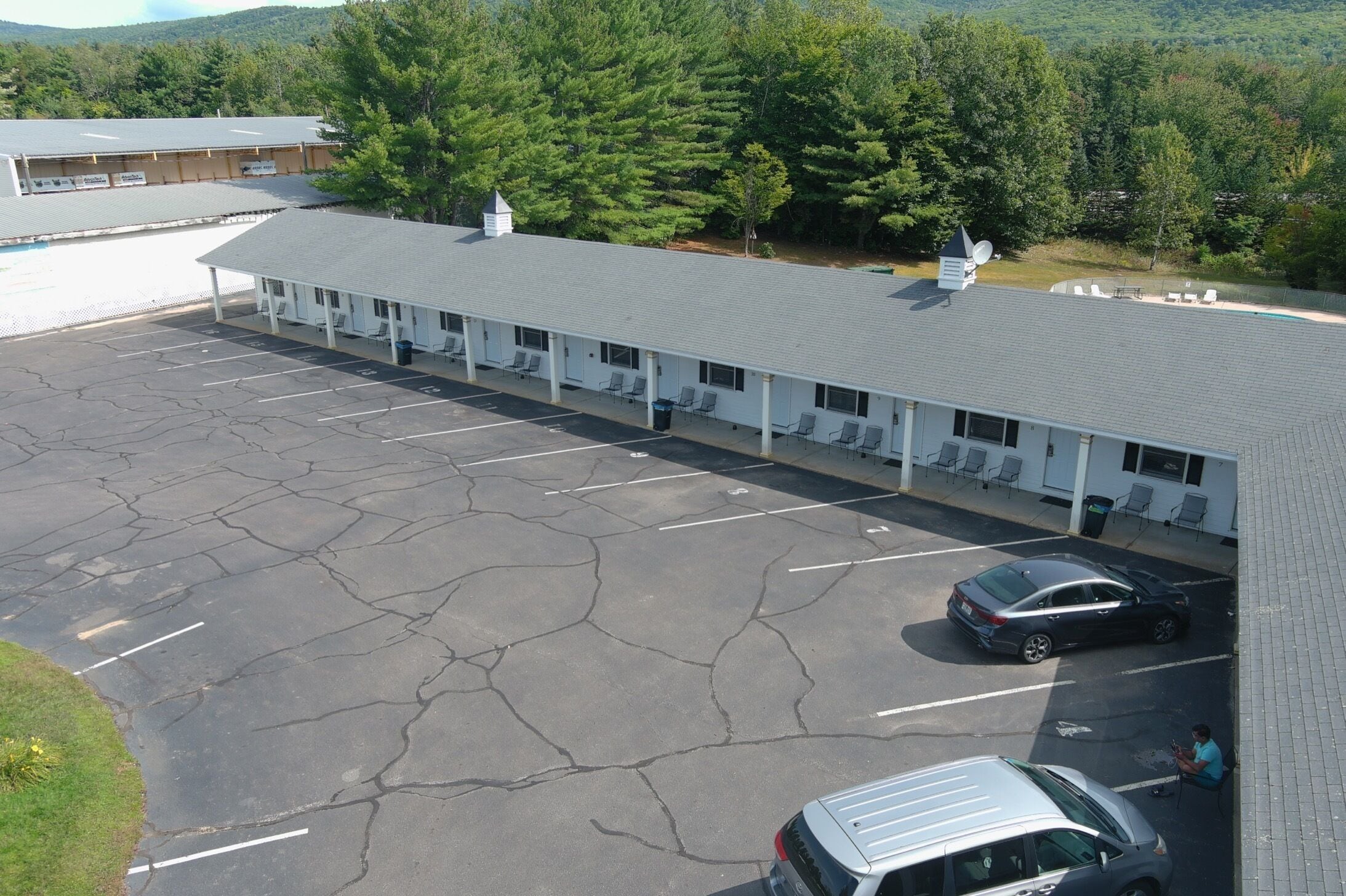 A motel with a row of rooms, a parking lot, and a view of a forested area.
