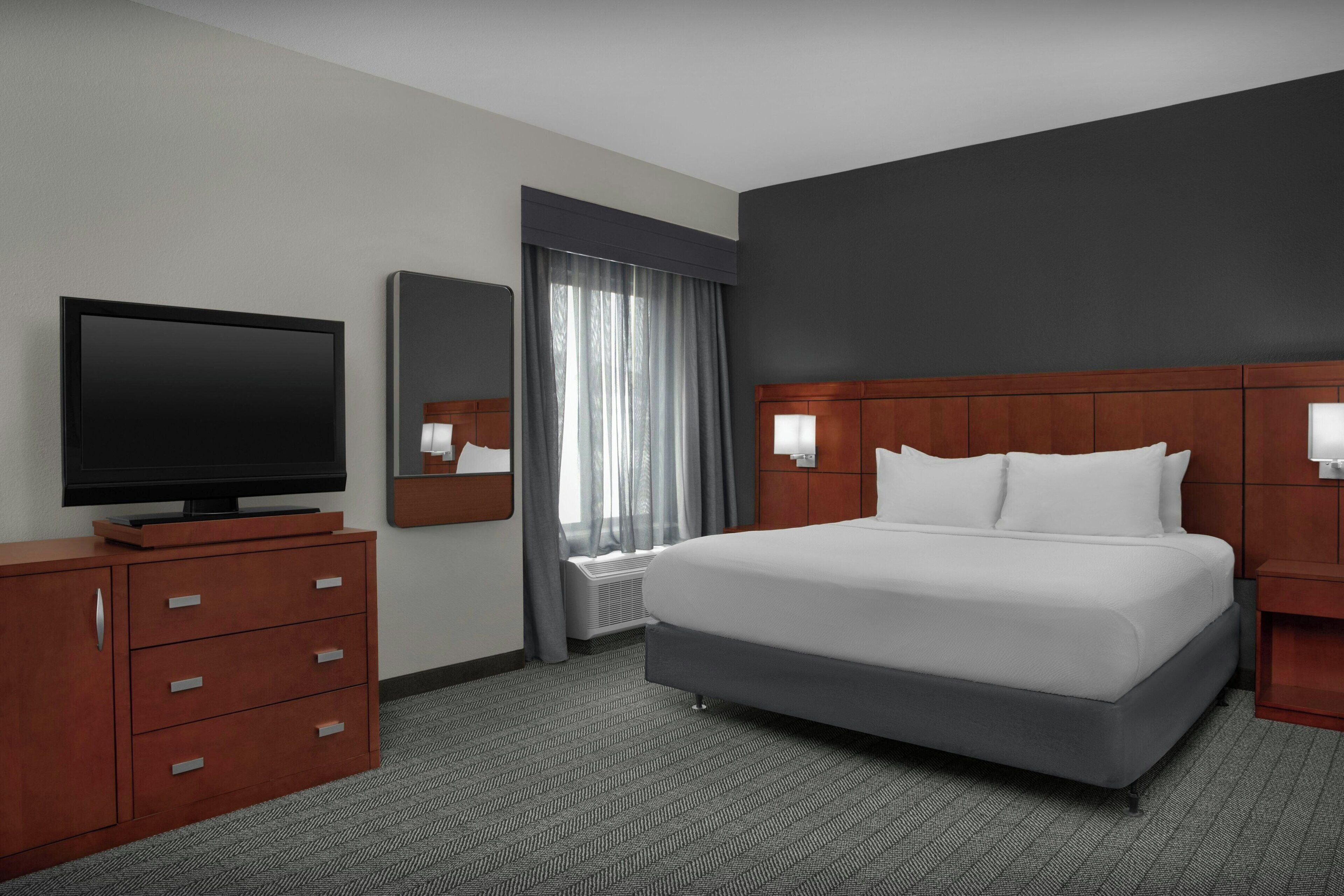 A hotel room with a bed, a television, a dresser, a mirror, and a window with curtains.
