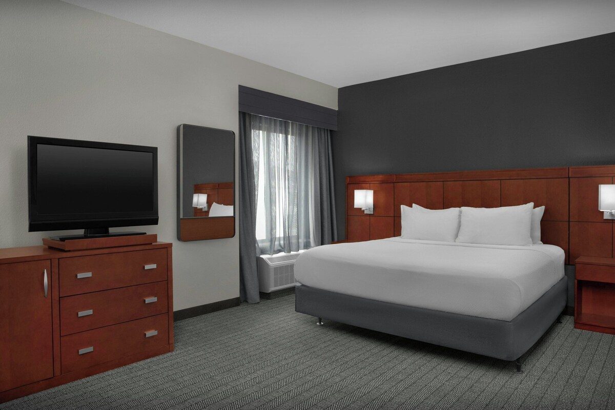 Habitación de hotel con cama, televisión, cómoda, espejo y ventana con cortinas.