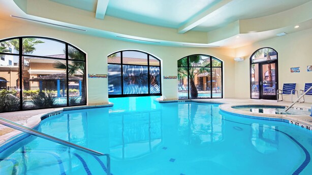 A large indoor swimming pool with clear blue water, surrounded by large arched windows offering views of palm trees and a building. - Courtyard by Marriott San Antonio SeaWorld - Westover Hills (San Antonio)