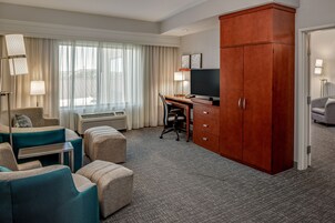 A hotel room with a desk, chair, and two sofas. There is a wardrobe, a television, and a lamp.