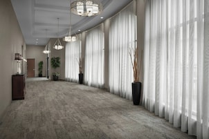 A hotel corridor with a carpeted floor, large windows with sheer curtains, and multiple hanging chandeliers.