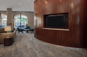 A modern lobby with a wooden reception desk, a flat-screen TV, comfortable seating, and large windows offering a view of the outdoors.