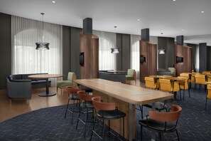 A modern hotel lobby with a central bar area, seating arrangements, and large windows with curtains.