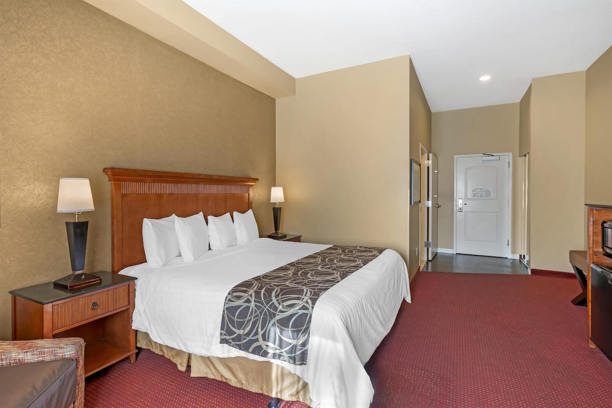 A hotel room with a large bed, two bedside tables with lamps, a wooden headboard, a red carpet, and a door leading to a hallway.