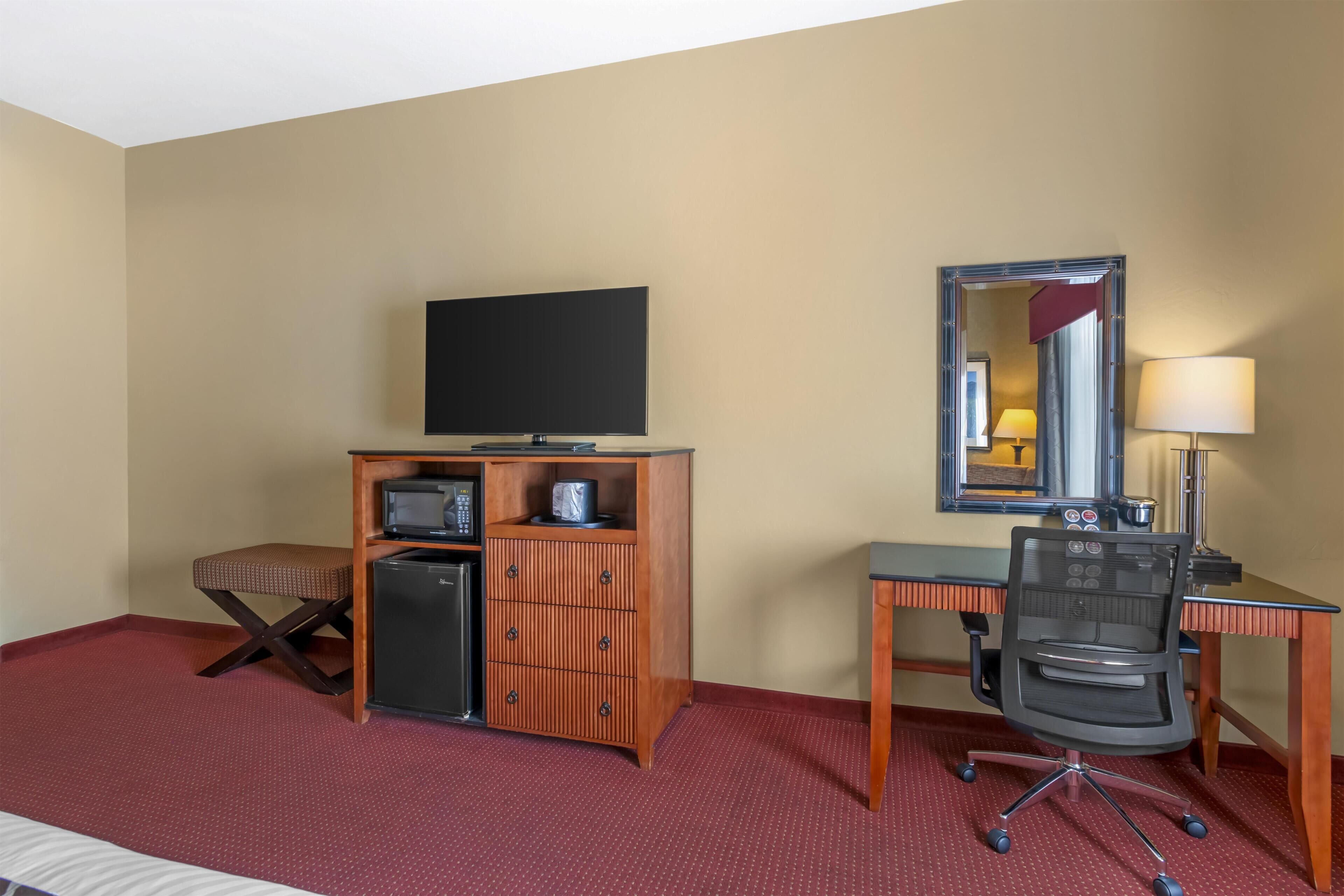 A hotel room with a bed, a desk with a computer, a television, a small refrigerator, a microwave, a chair, and a mirror.