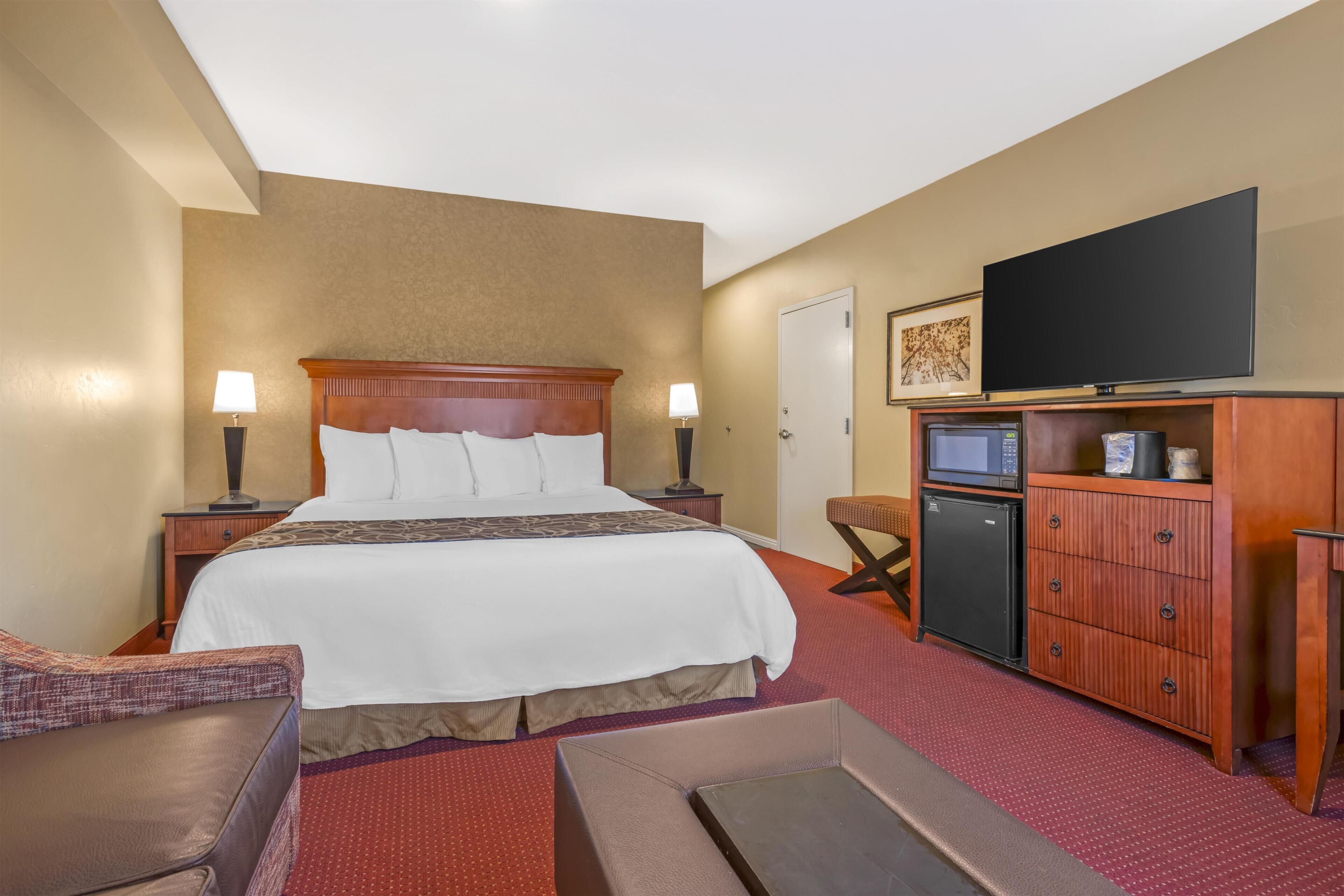 Una habitación de hotel con una cama grande, televisión, escritorio con computadora y un refrigerador pequeño.