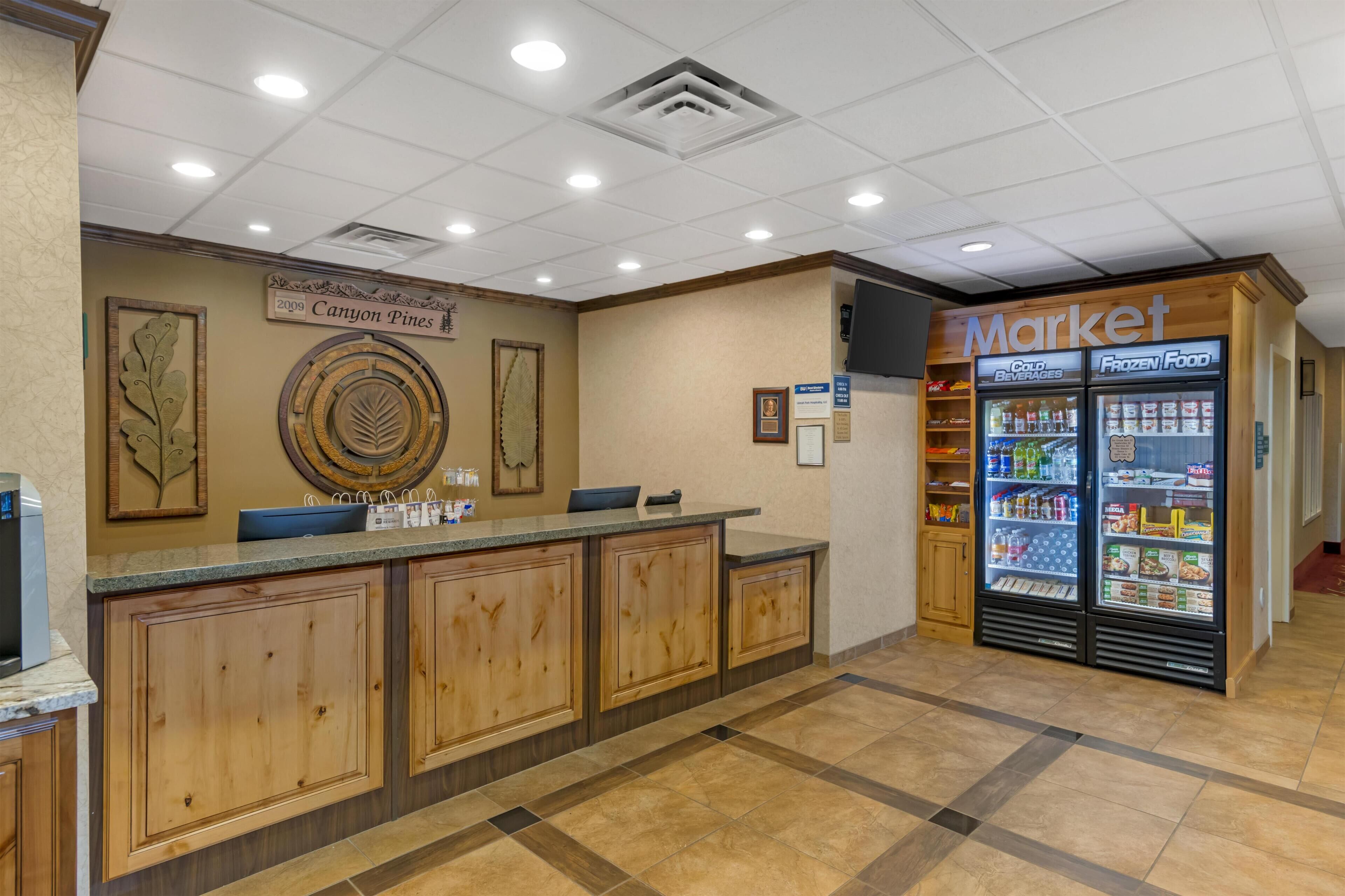 A reception area with a wooden reception desk, a wall-mounted sign reading "Canyon Pines," and a "Market" refrigerator stocked with beverages and frozen food.
