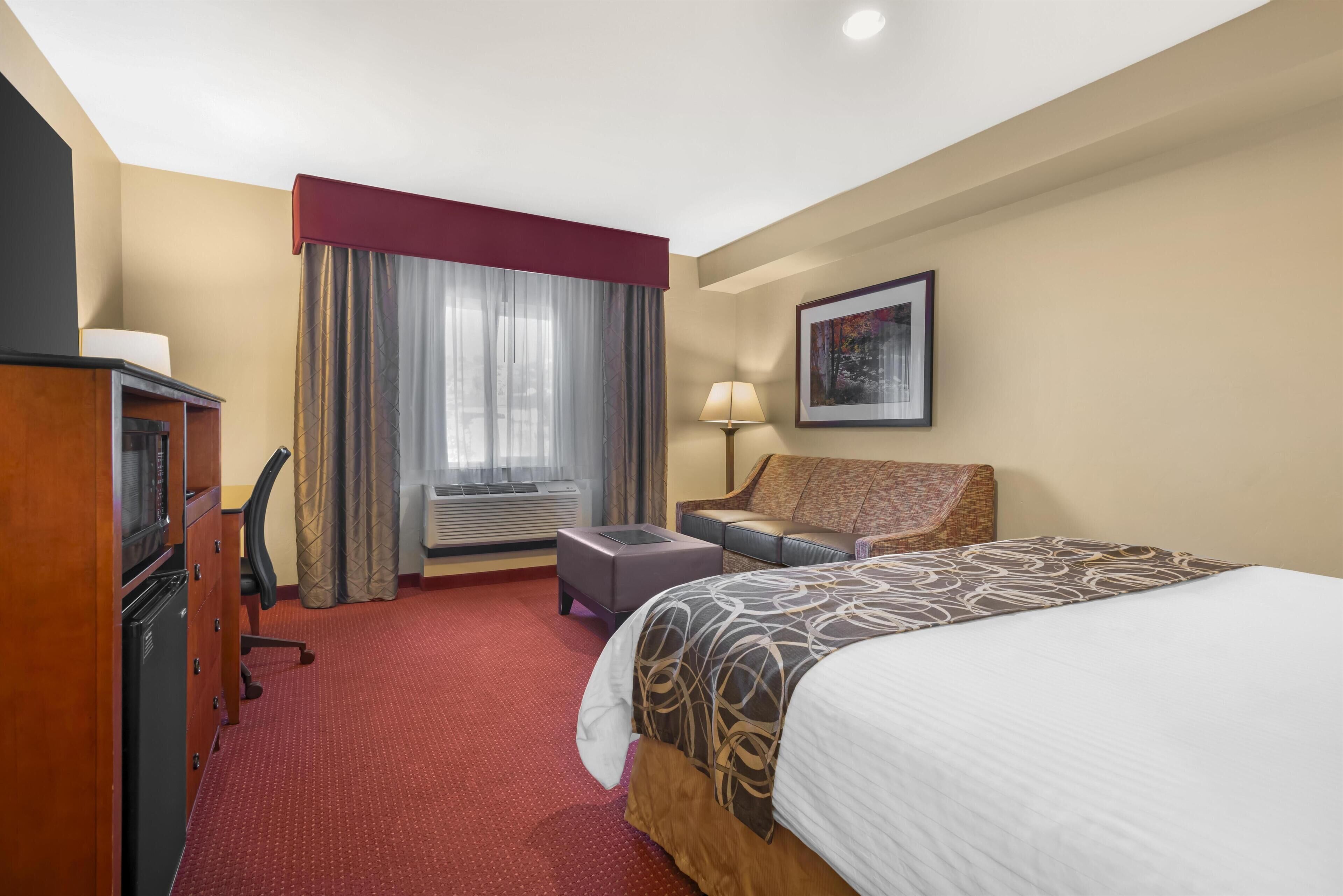 Une chambre d’hôtel avec un lit, un bureau, une chaise, une lampe, un tableau au mur et une fen être avec des rideaux.