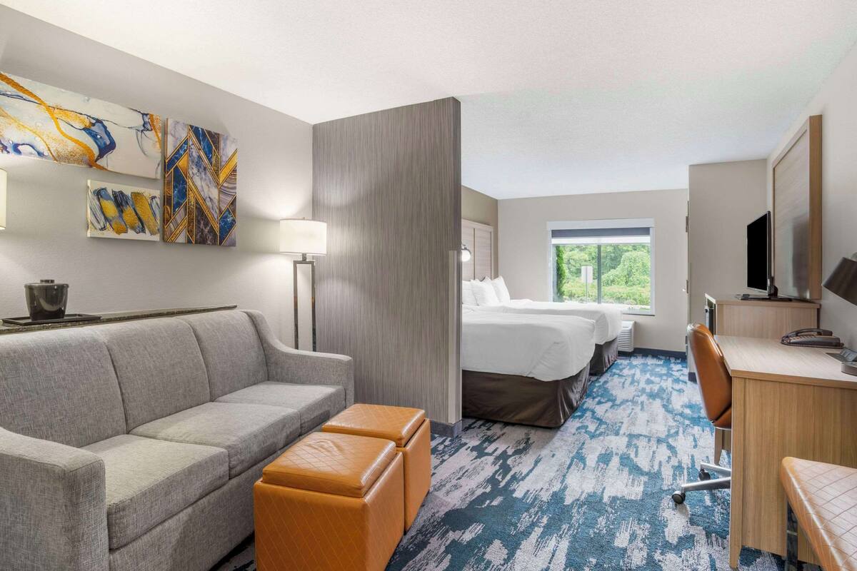 A hotel room with a sofa, two ottomans, a desk, a bed, and artwork on the wall.