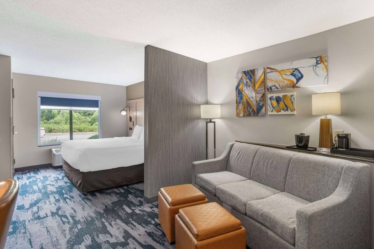 A hotel room with a bed, a gray sofa, two ottomans, a window with a view, and abstract artwork on the wall.