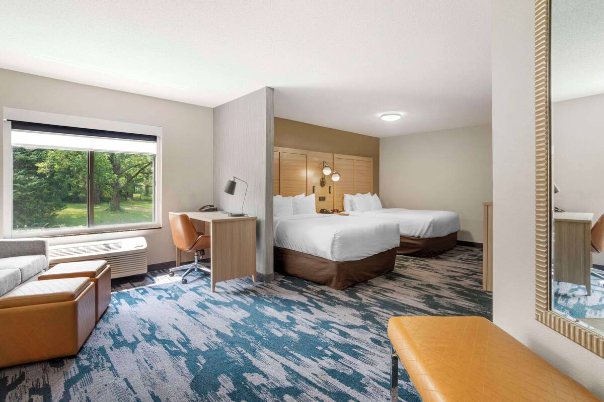 A hotel room with a large bed, a desk, a chair, a bench, and a large window with a view of trees.