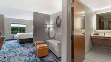 A hotel room with a bed, a desk, a chair, a sofa, a lamp, and a bathroom with a mirror and sink.