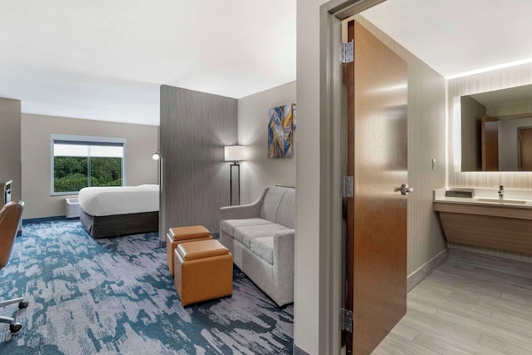 Habitación de hotel con cama, escritorio, silla, sofá, lámpara y baño con espejo y lavamanos.