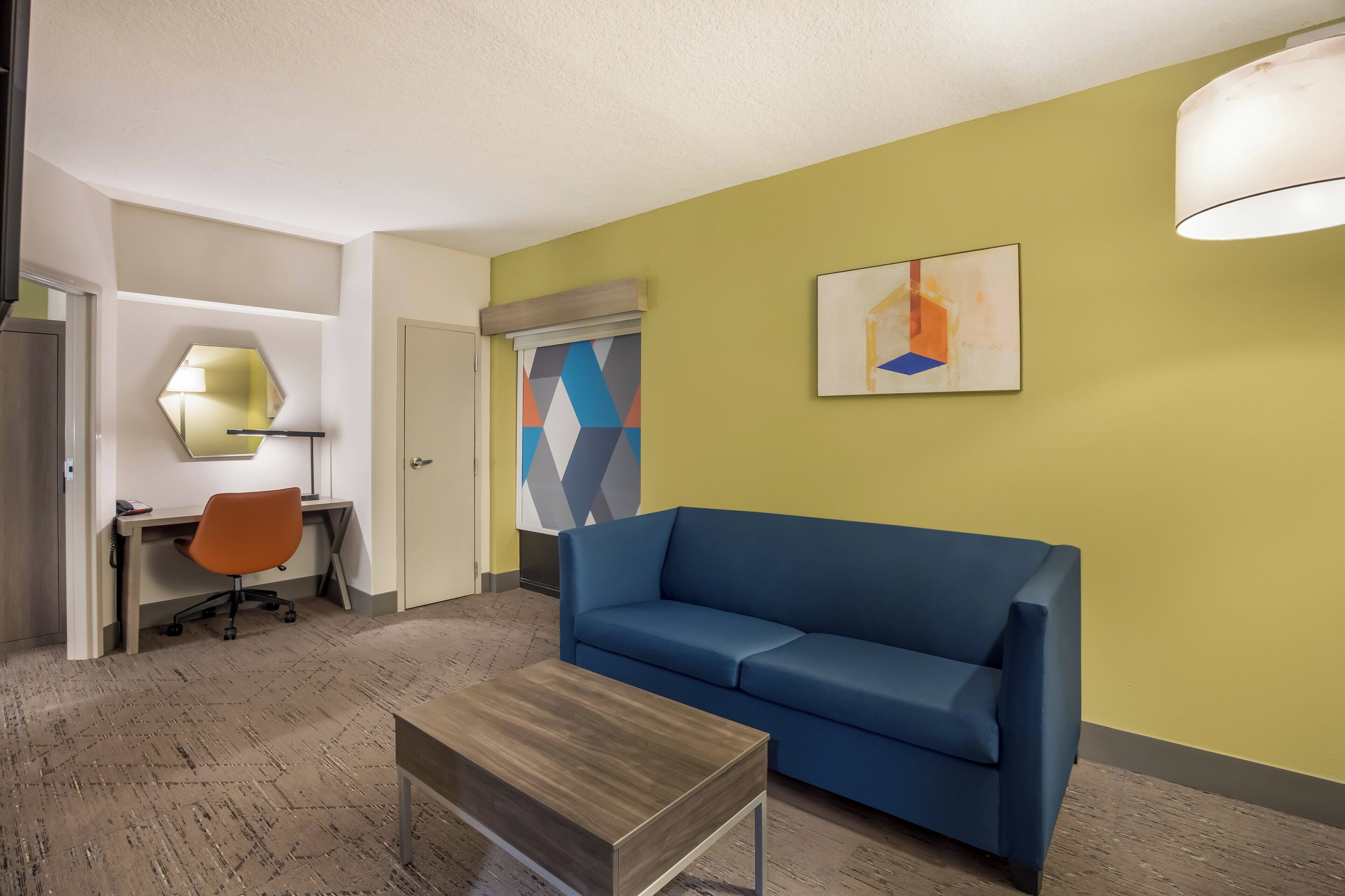 A modern hotel room with a blue sofa, a wooden coffee table, a desk with a chair, a lamp, and a geometric painting on the wall.