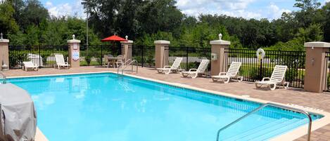 A swimming pool with lounge chairs and a covered area, surrounded by a fence and greenery.