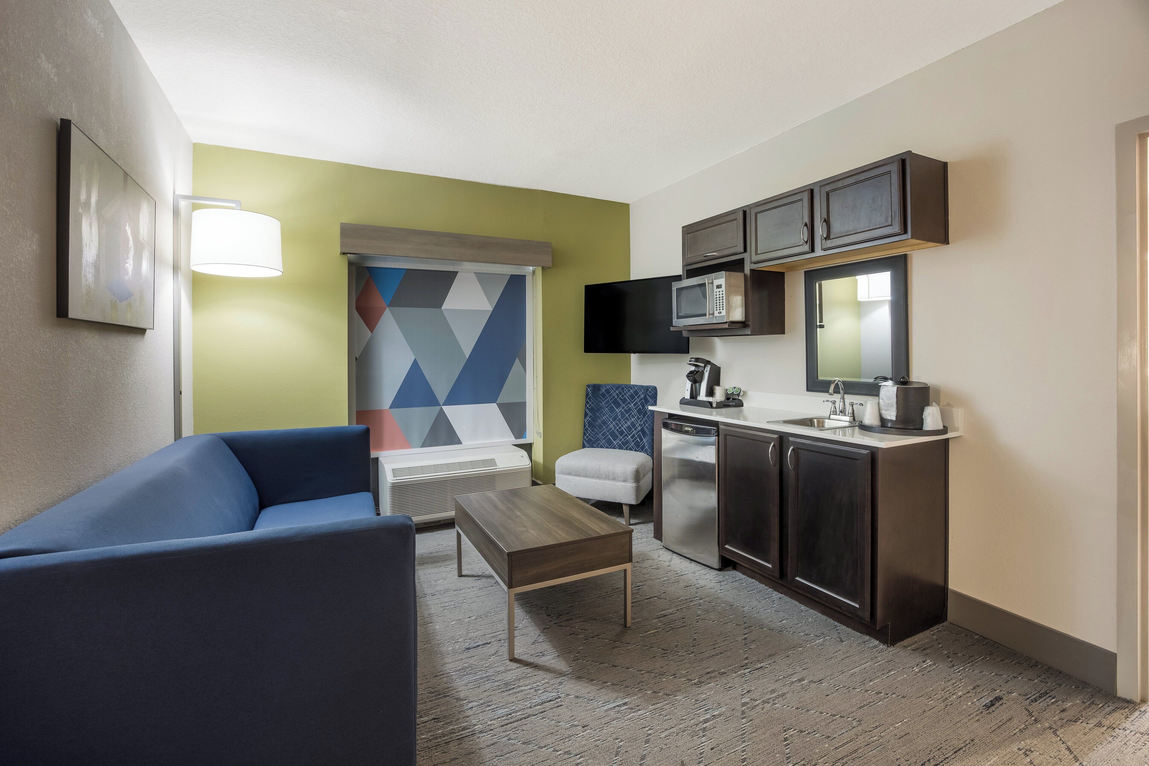 Habitación de hotel compacta con kitchenette, sofá, mesita pequeña y pared con llamativo estampado geométrico.