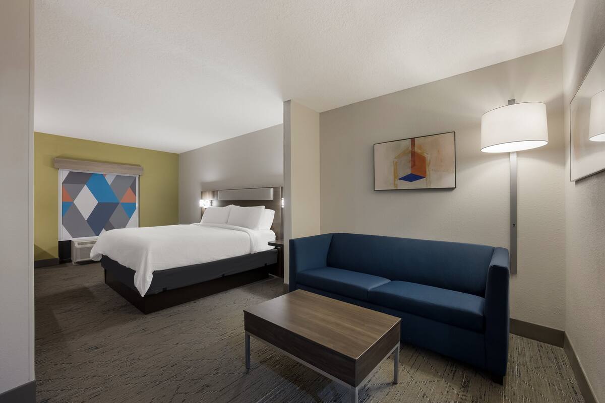 A hotel room with a bed, a blue sofa, a wooden coffee table, and a painting on the wall.