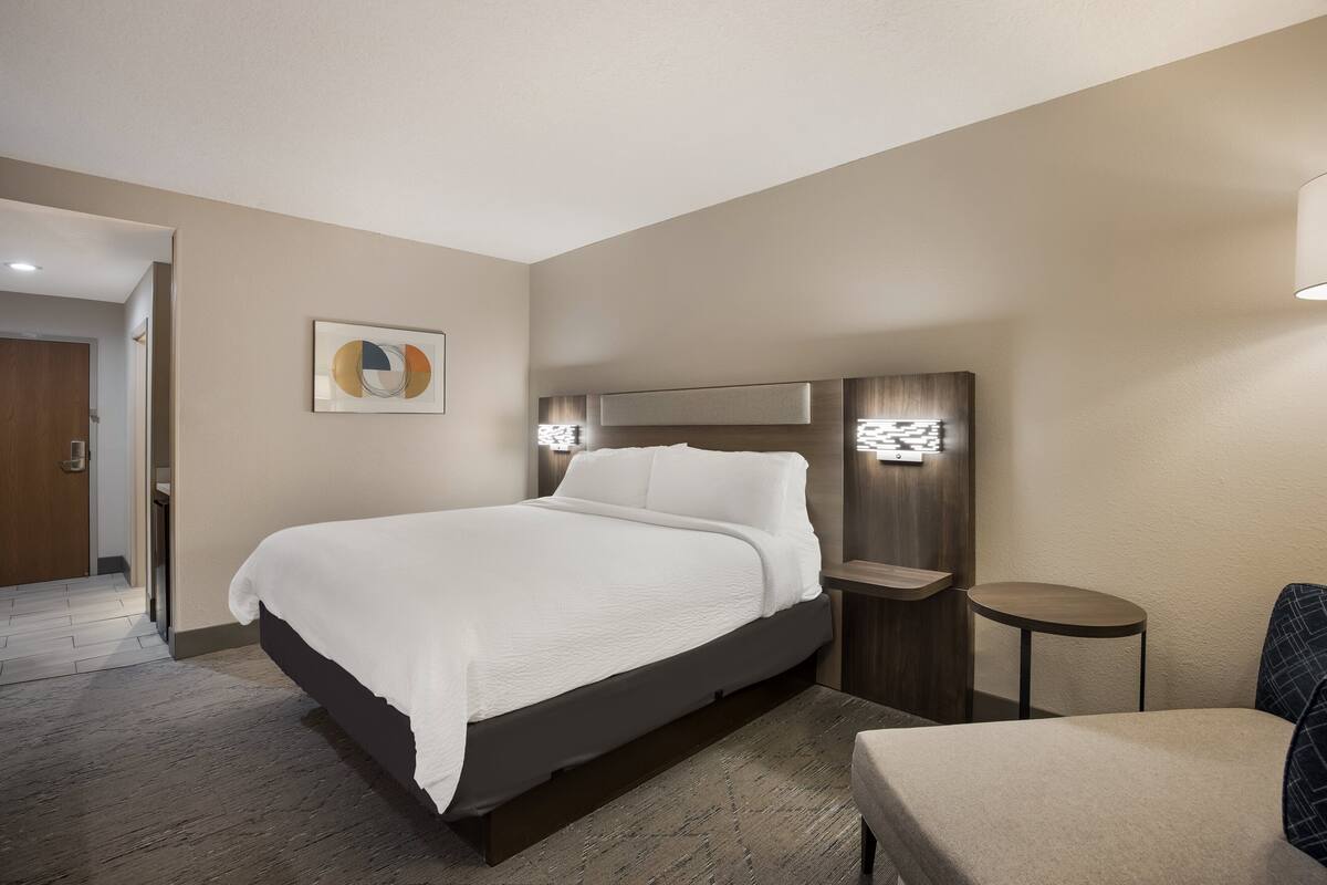 A modern hotel room with a large bed, a bedside table, a small round table, and a wall-mounted light fixture.
