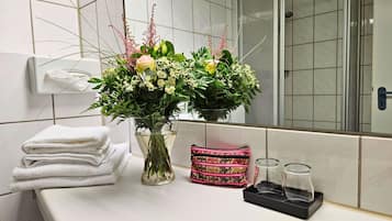 Un baño con espejo, un jarrón de flores, toallas dobladas, un bolso estampado y dos recipientes de vidrio vacíos sobre una encimera.