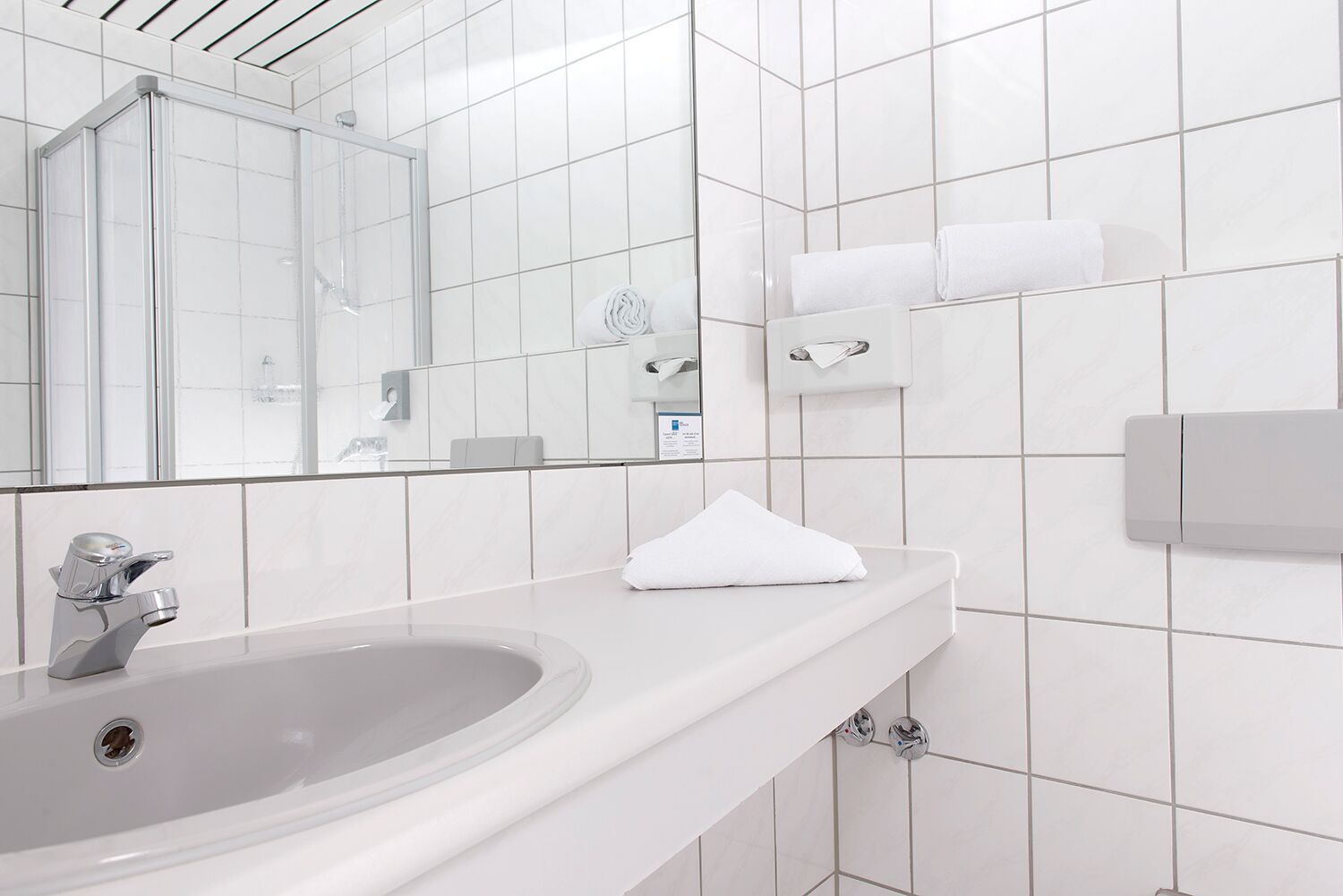 A modern bathroom with a white sink, a glass shower enclosure, a mirror, and a towel dispenser.