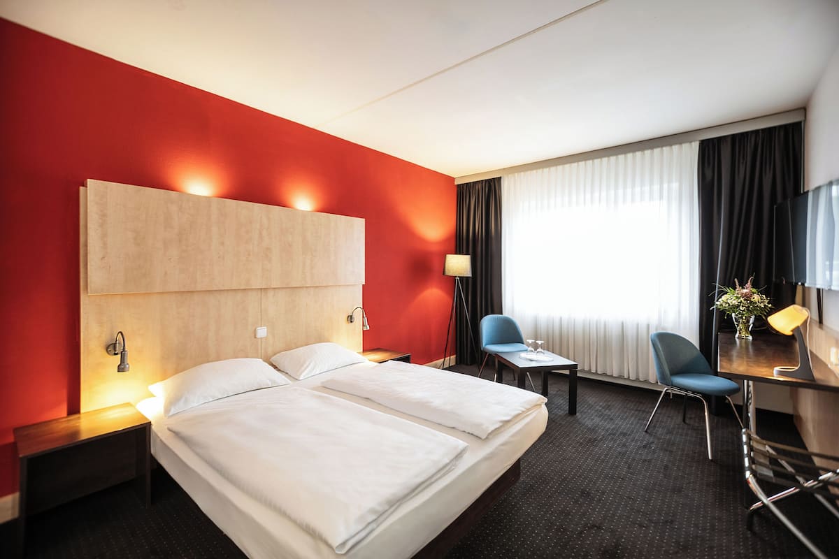 A hotel room with a large bed, a desk with a chair, a lamp, and a window with curtains.