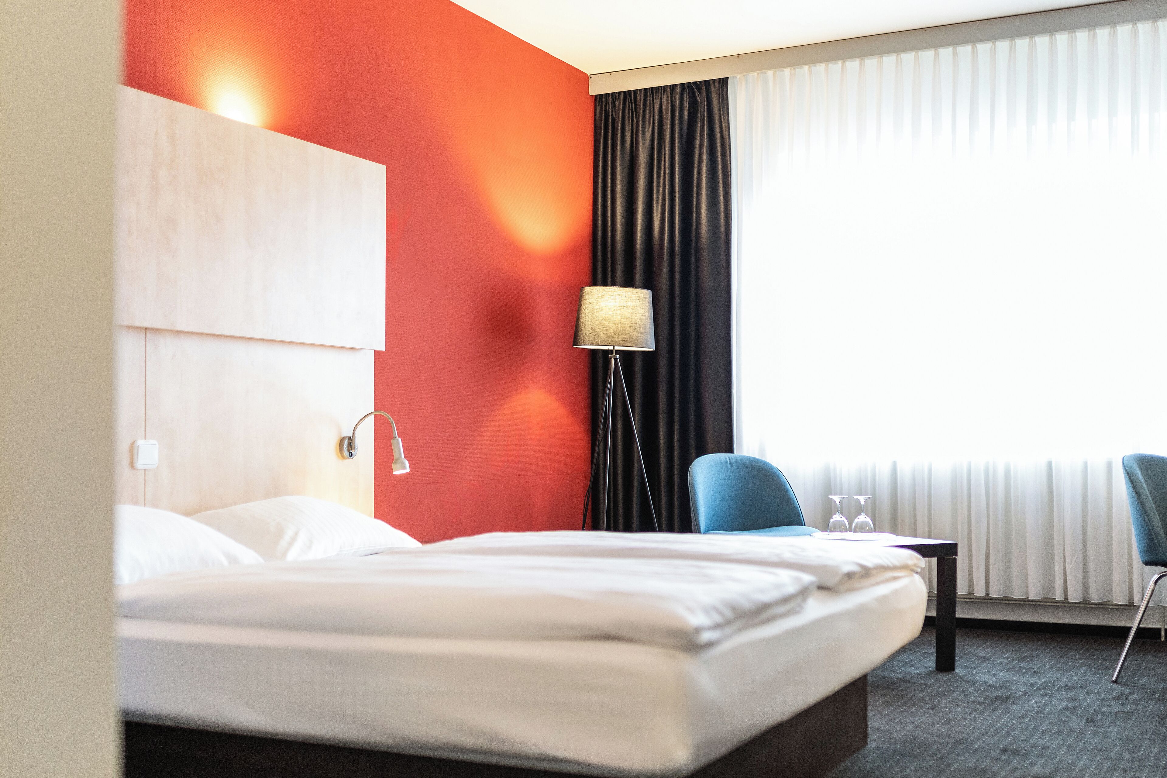 A hotel room with a bed, a desk, two chairs, a lamp, and a window with curtains.