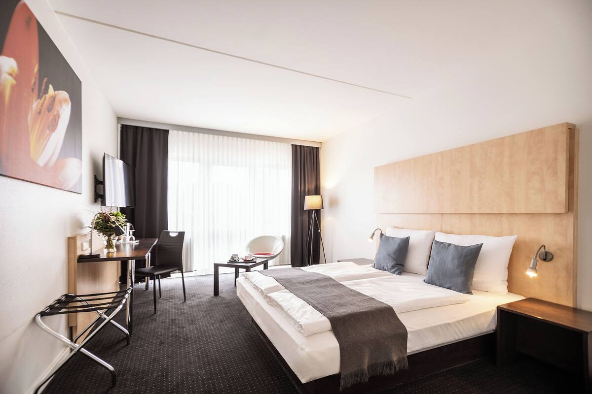 A hotel room with a bed, bedside table, lamp, desk, chair, and a large window with curtains.