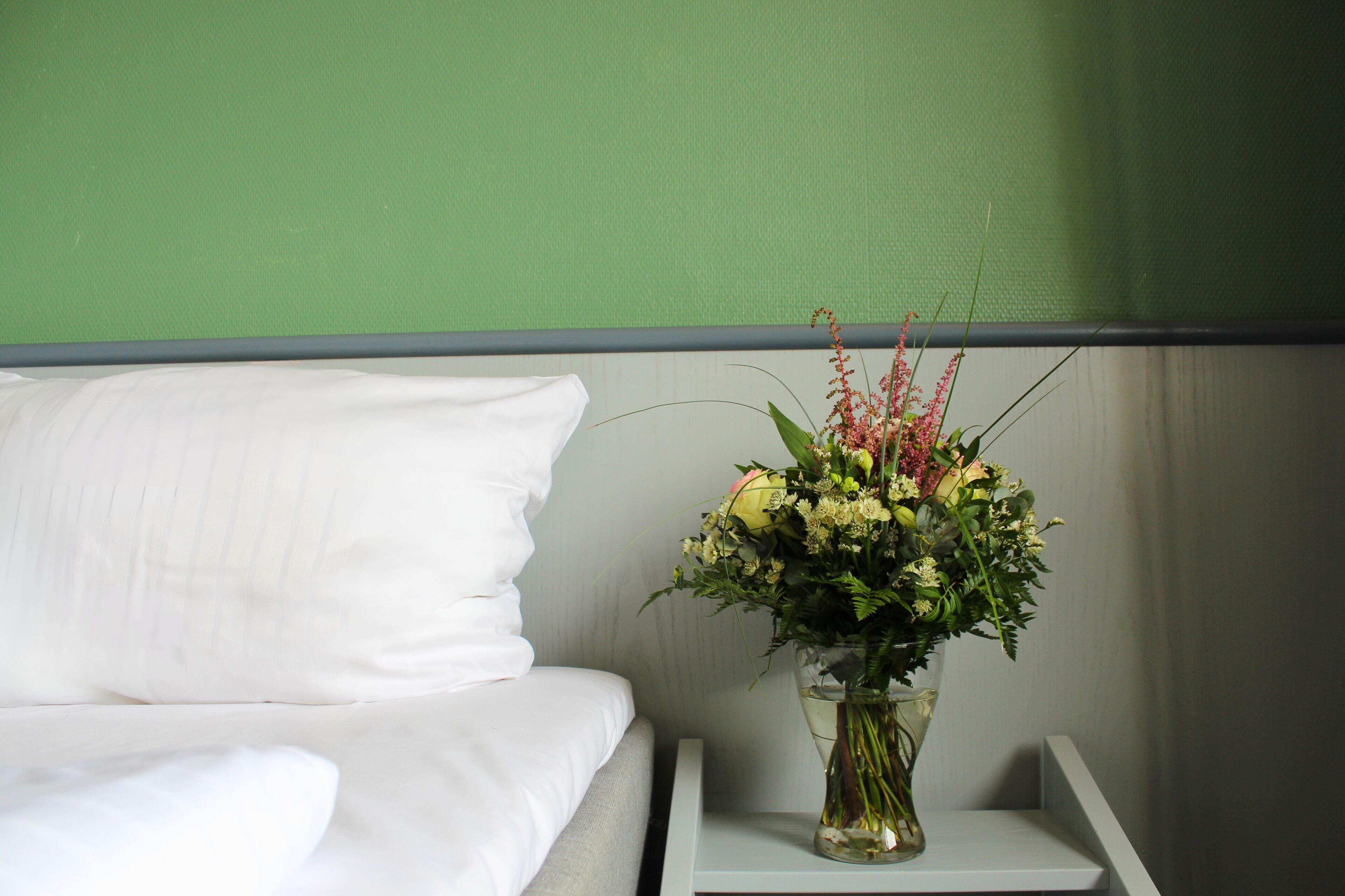 Una cama con sábanas blancas y una mesita de noche con un florero transparente lleno de flores.