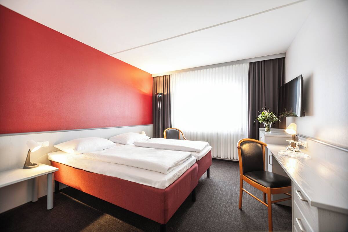 A hotel room with a red accent wall, a bed with white linens, a desk with a chair, a flat-screen TV, and a window with curtains.