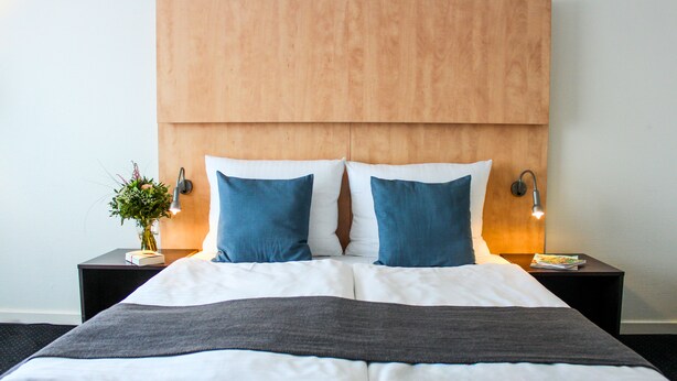 Cama bem arrumada com roupa de cama branca e azul, duas mesas de cabeceira com abajures e uma cabeceira de madeira.