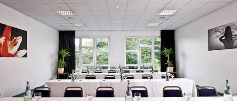 Un salón de conferencias con varias mesas, sillas y ventanas amplias que ofrecen vistas al follaje exterior.
