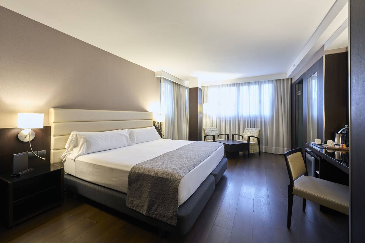 Une chambre d’hôtel avec un grand lit, une table de chevet, une lampe, un bureau, une chaise et des rideaux.