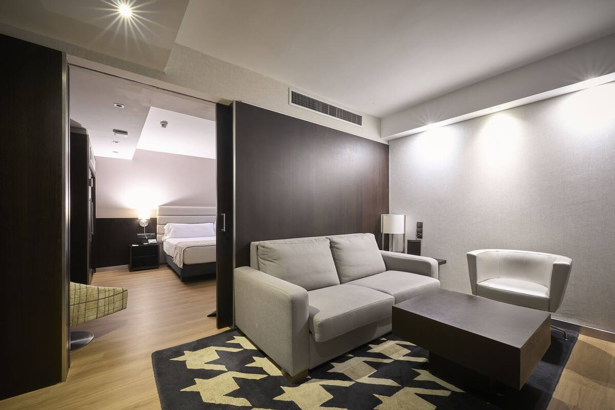 Une chambre d’hôtel moderne avec un canapé gris, une petite table, un fauteuil blanc et un lit en arrière-plan.