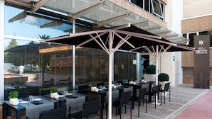 Un área de comedor al aire libre con mesas y sillas, un cerramiento de vidrio y un cartel para el Hotel Talonia Rigolett.