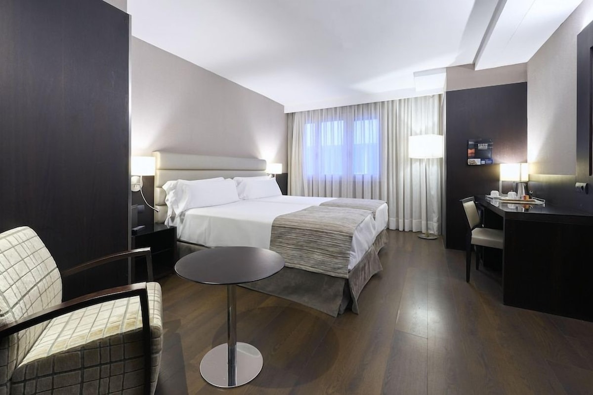 Habitación de hotel con cama, escritorio, silla, una mesa redonda y un ventanal con cortinas.