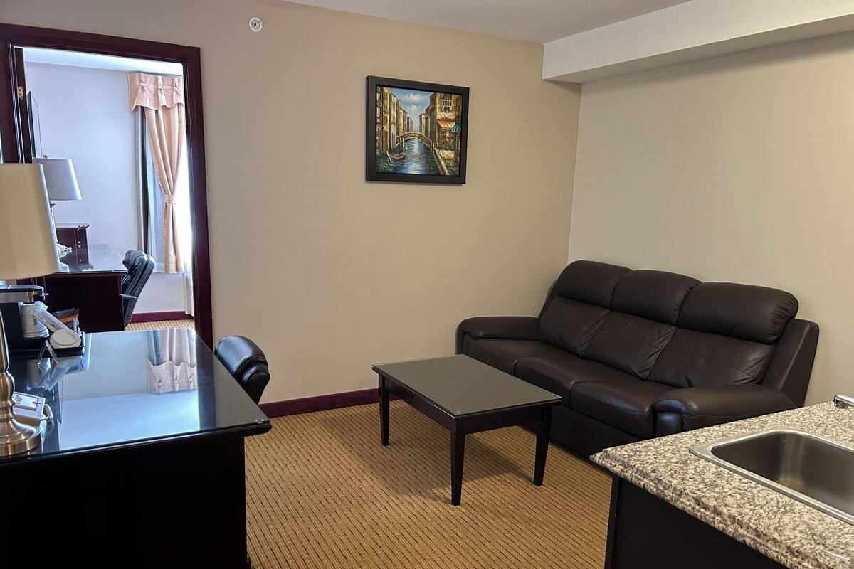 A hotel room with a leather sofa, a small table, a desk with a lamp, a painting on the wall, and a kitchen area with a sink.