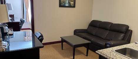 Habitación de hotel con sofá de cuero, una mesita, un escritorio con lámpara, un cuadro en la pared y una zona de cocina con fregadero.