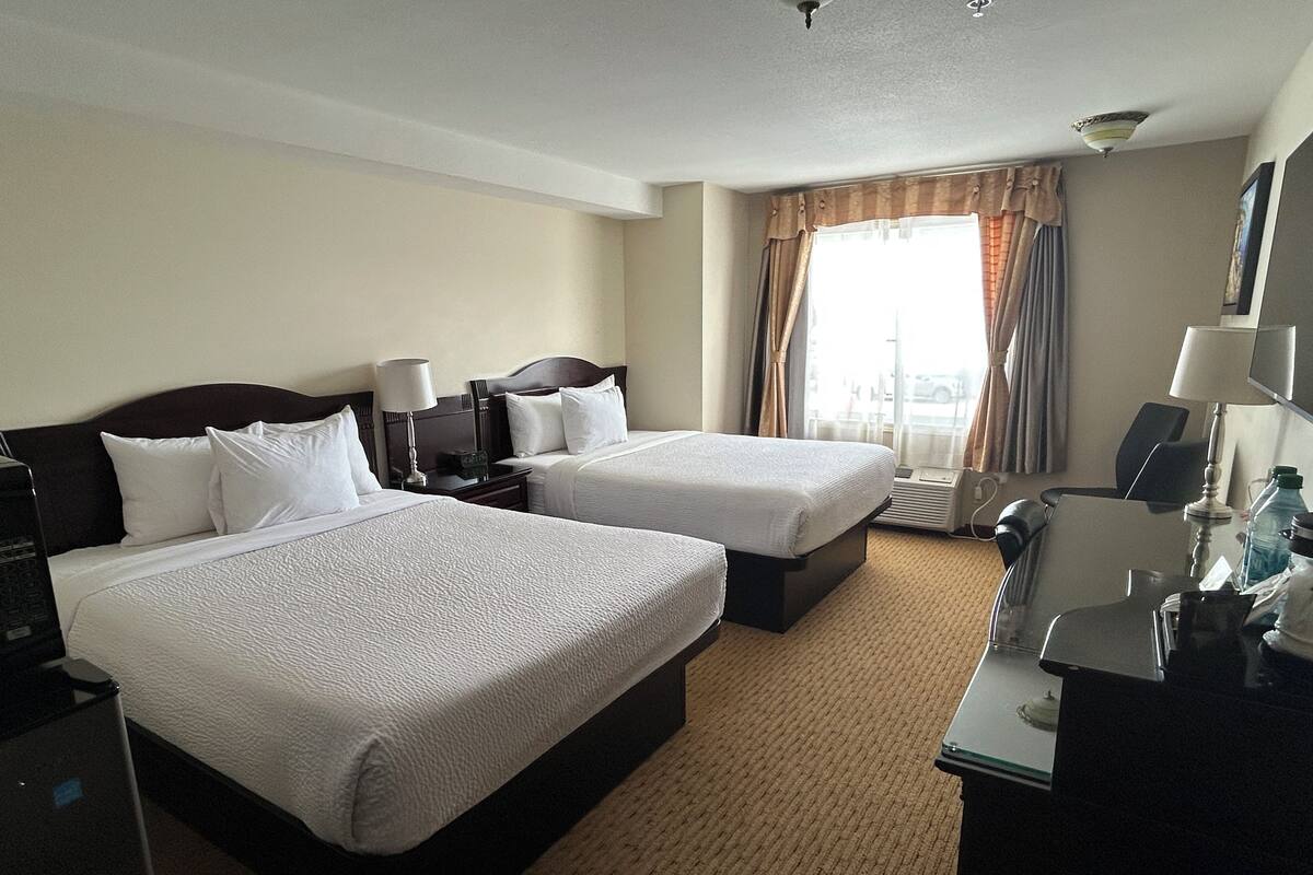 A hotel room with two beds, a desk, a chair, a lamp, and a window with curtains.