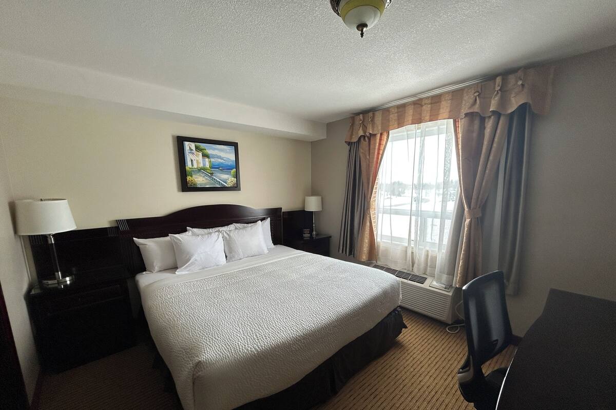 A hotel room with a bed, a desk, a chair, a lamp, a painting on the wall, and a window with curtains.