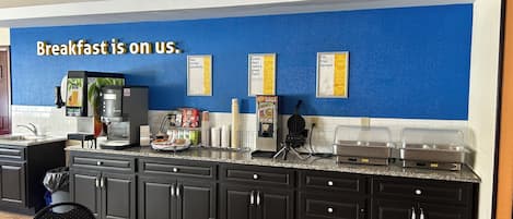 A breakfast area with a blue wall, coffee machines, a waffle maker, and a sign that says 'Breakfast is on us.'