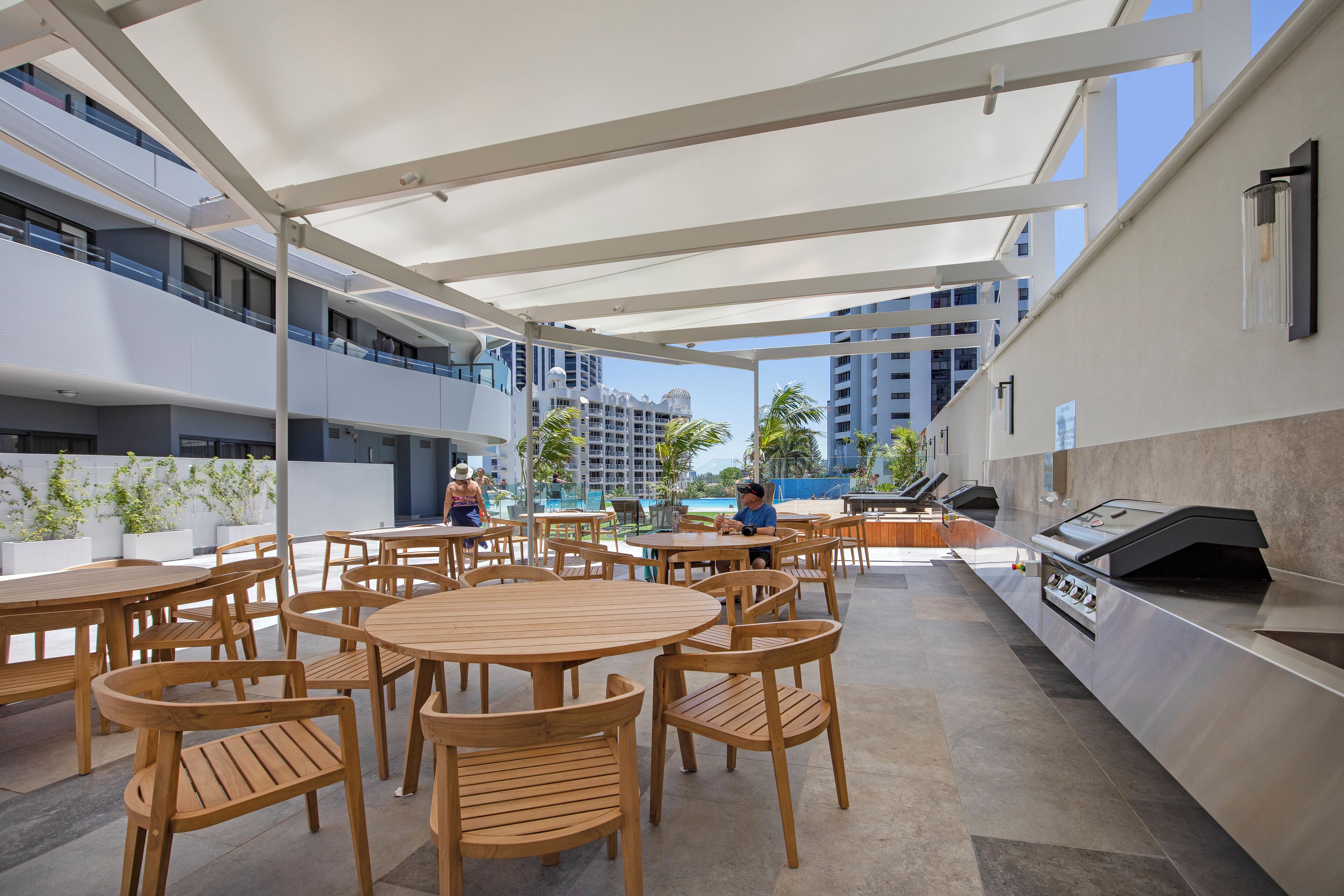 An outdoor dining area with wooden tables and chairs, a grill station, and a view of a building with balconies and palm trees.