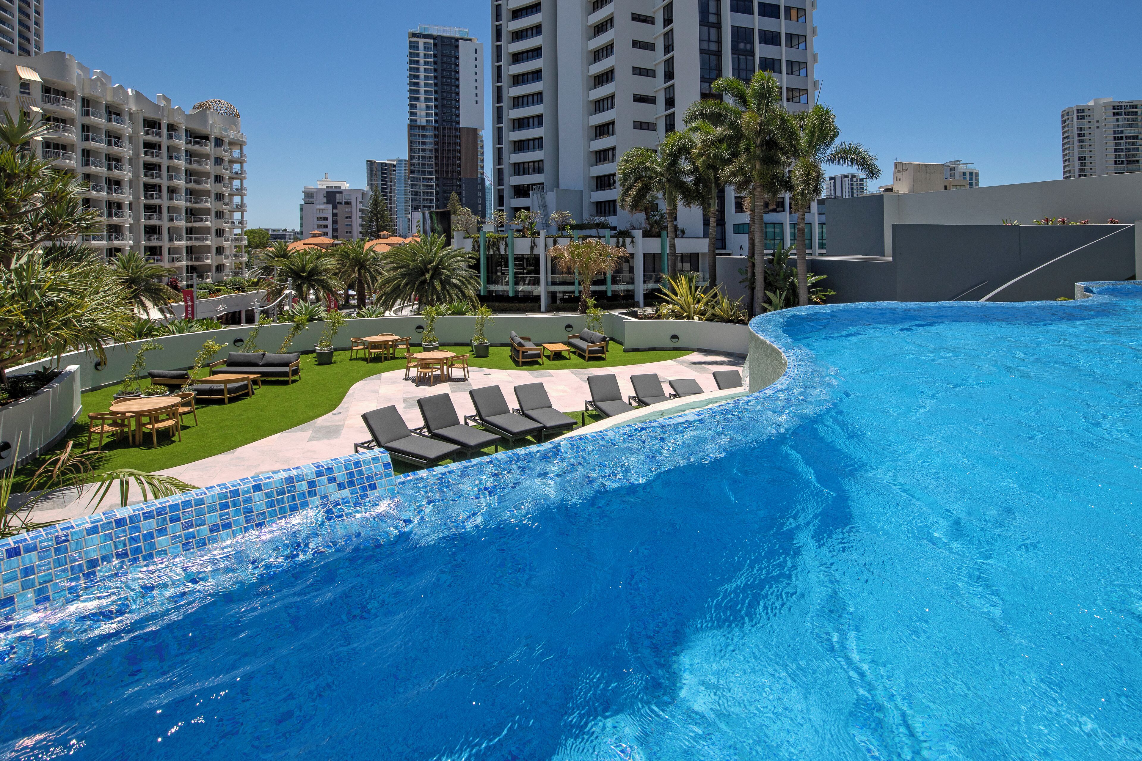 A rooftop pool area with lounge chairs and palm trees, surrounded by high-rise buildings.