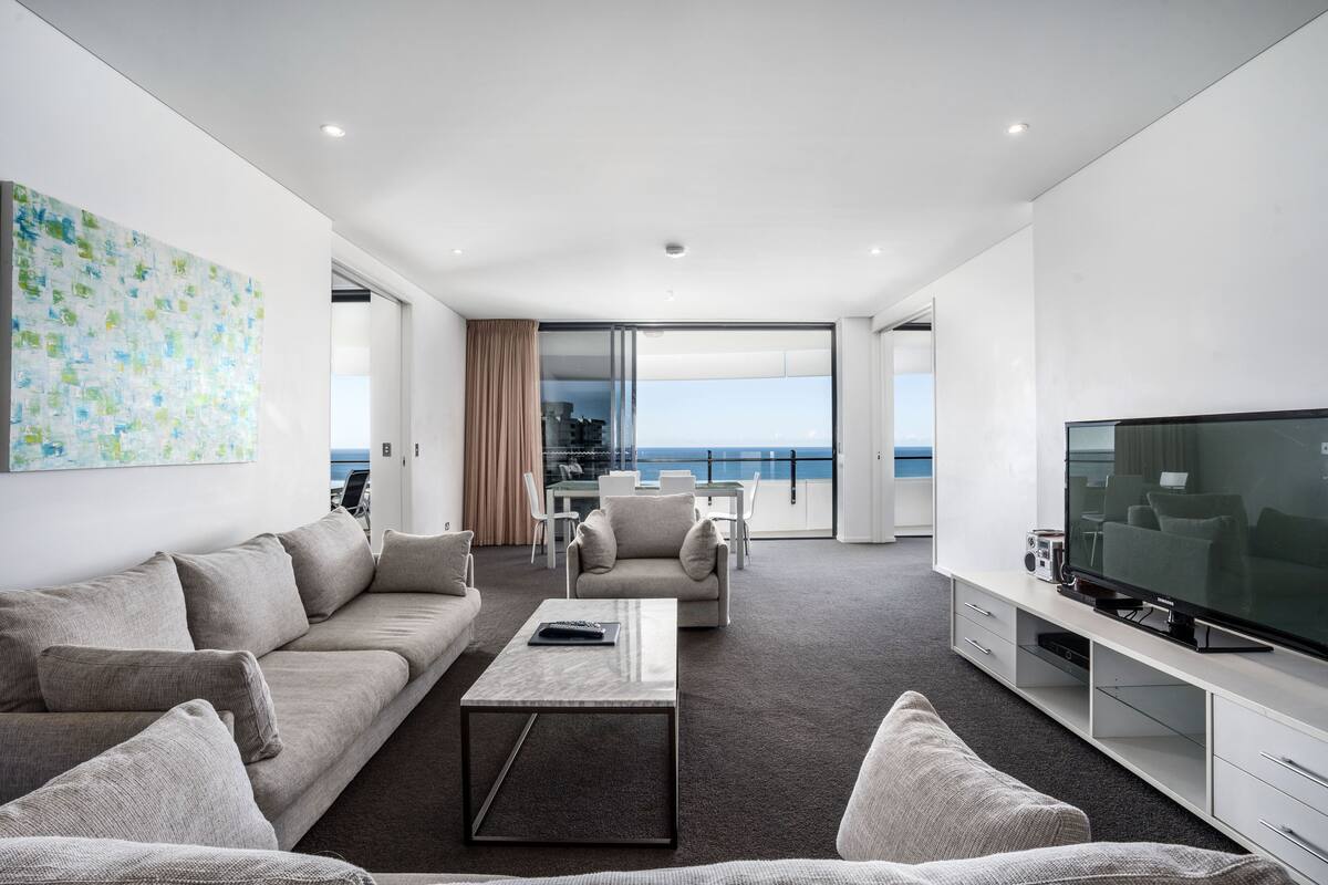 Una sala moderna con un televisor de pantalla plana grande, un sofá, una mesa de centro y un balcón con vista al mar.