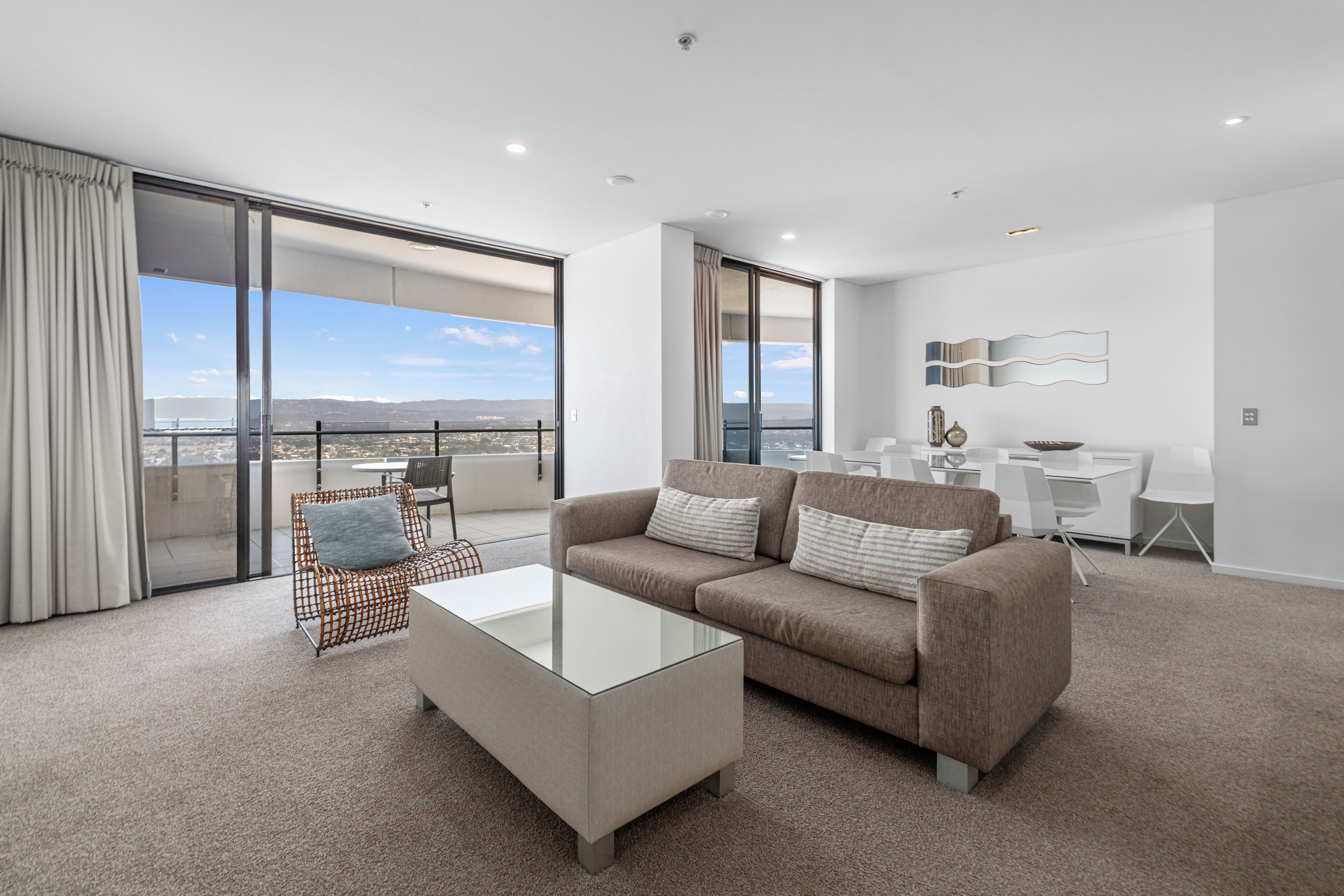 A modern living room with a glass coffee table, a sofa with cushions, a wicker chair, and a dining area with a white table and chairs.