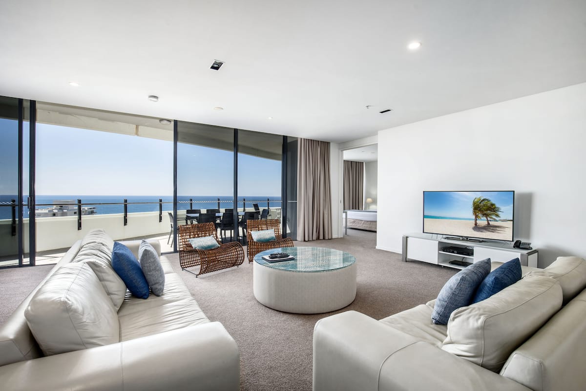 Una sala de estar moderna con un amplio ventanal con vistas al mar, una mesa de centro redonda de vidrio y un televisor de pantalla plana sobre un soporte blanco.