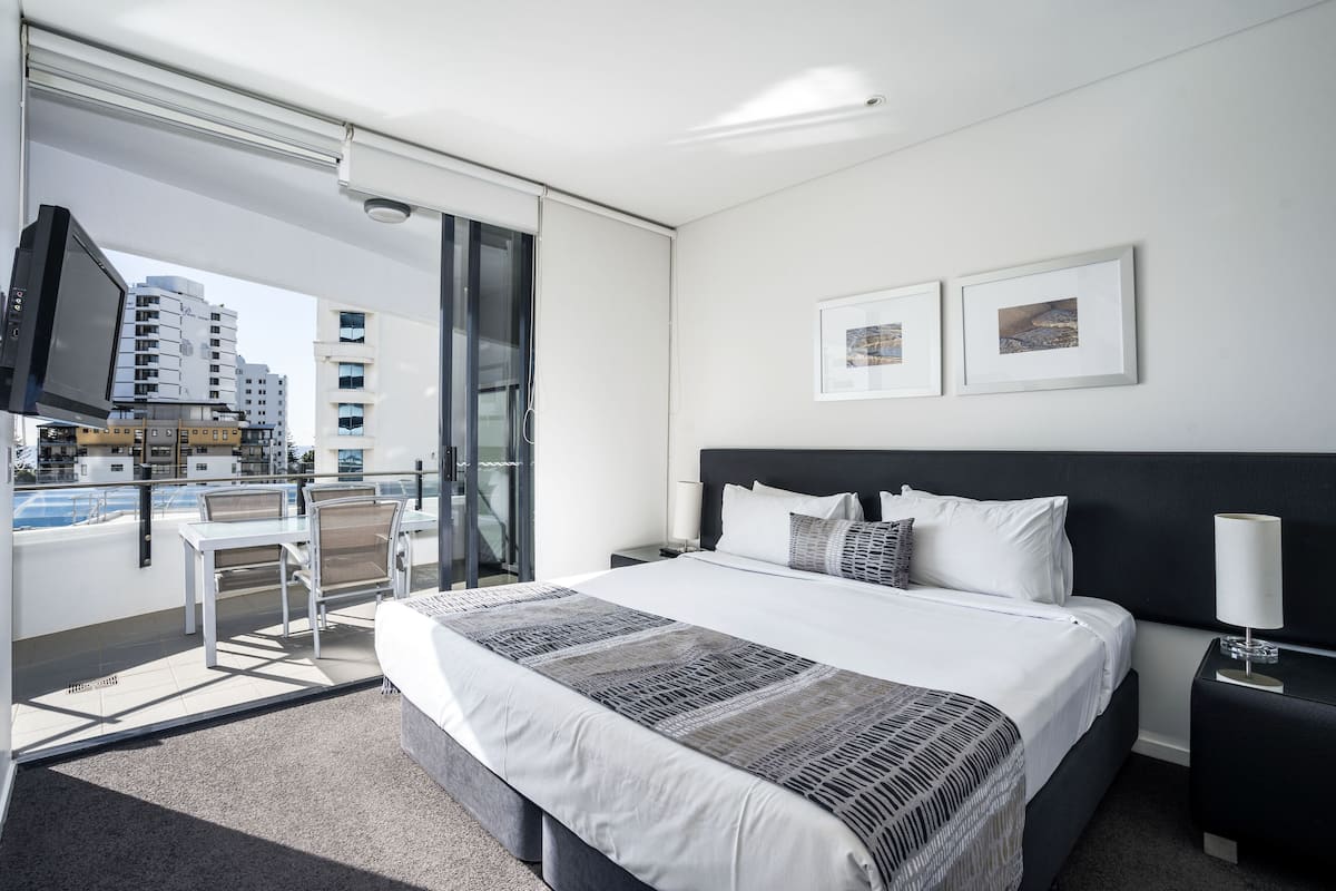 Una habitación de hotel moderna con una cama grande, un balcón con vistas a la ciudad, un televisor y una pequeña mesa con sillas.